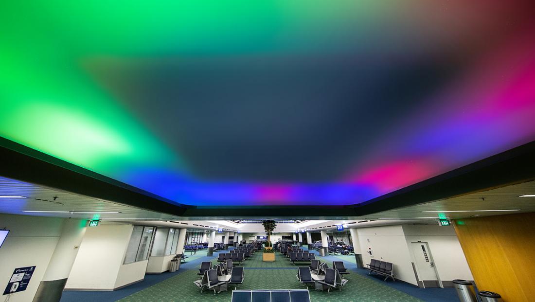 An airport gate lounge with its ceiling washed in green, blue and magenta light drifting the length of the concourse above empty seating.