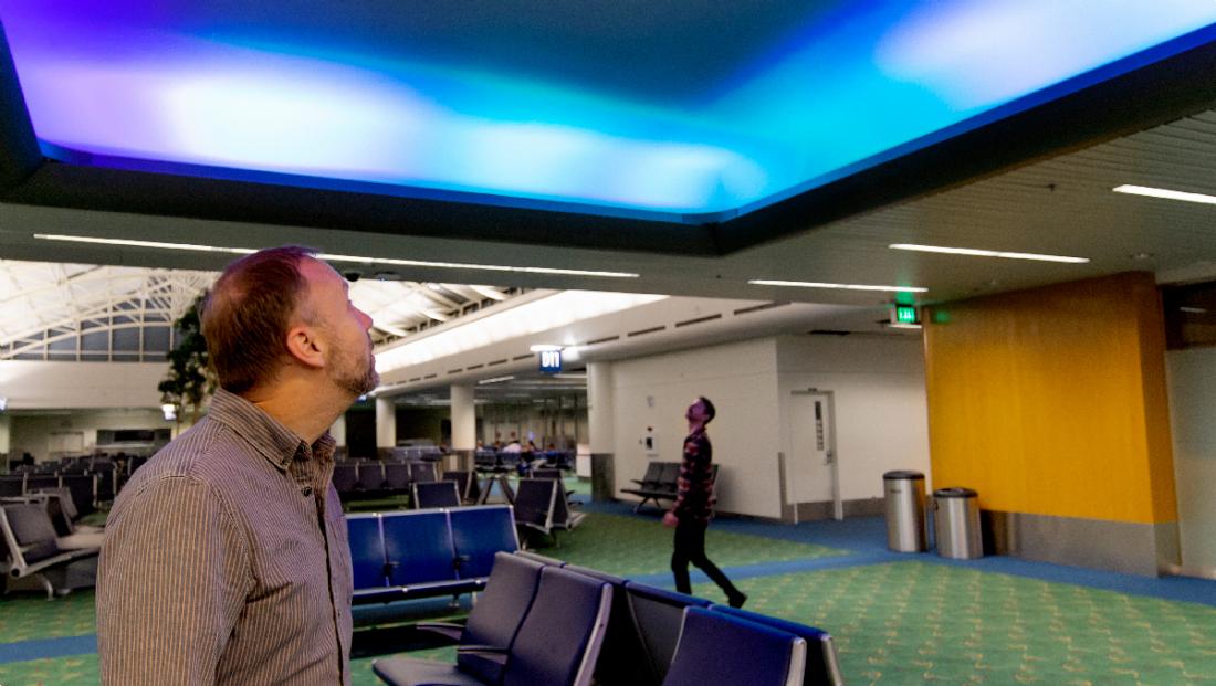 A traveller standing among the gate seating, looking up at the ceiling as it glows blue and violet.
