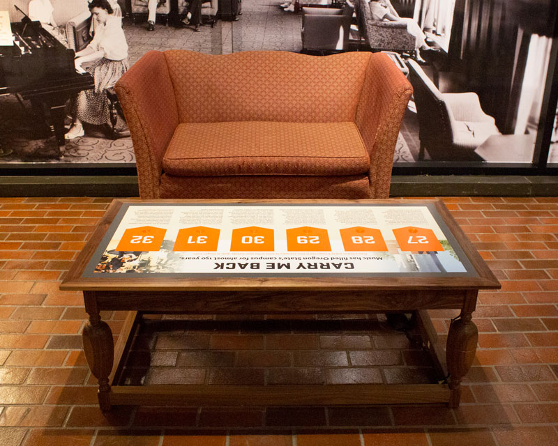 An armchair beside a wooden coffee table inlaid with a row of orange touch buttons, one labelled “Carry Me Back”, on a brick floor.