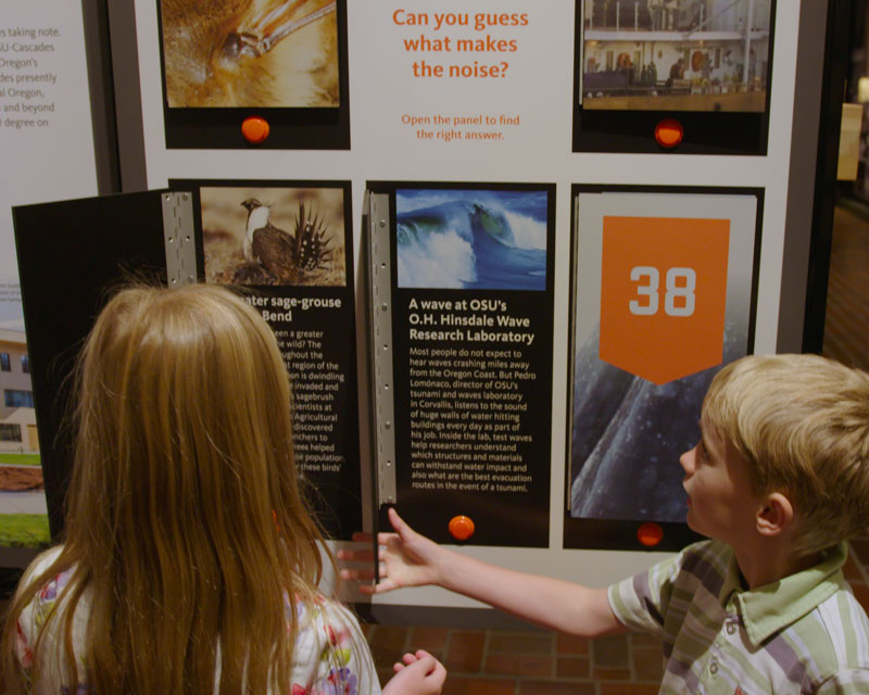 Two children at an exhibit panel headed “Can you guess what makes the noise?”, one pressing an orange button beside a card about OSU’s O.H. Hinsdale Wave Research Laboratory.