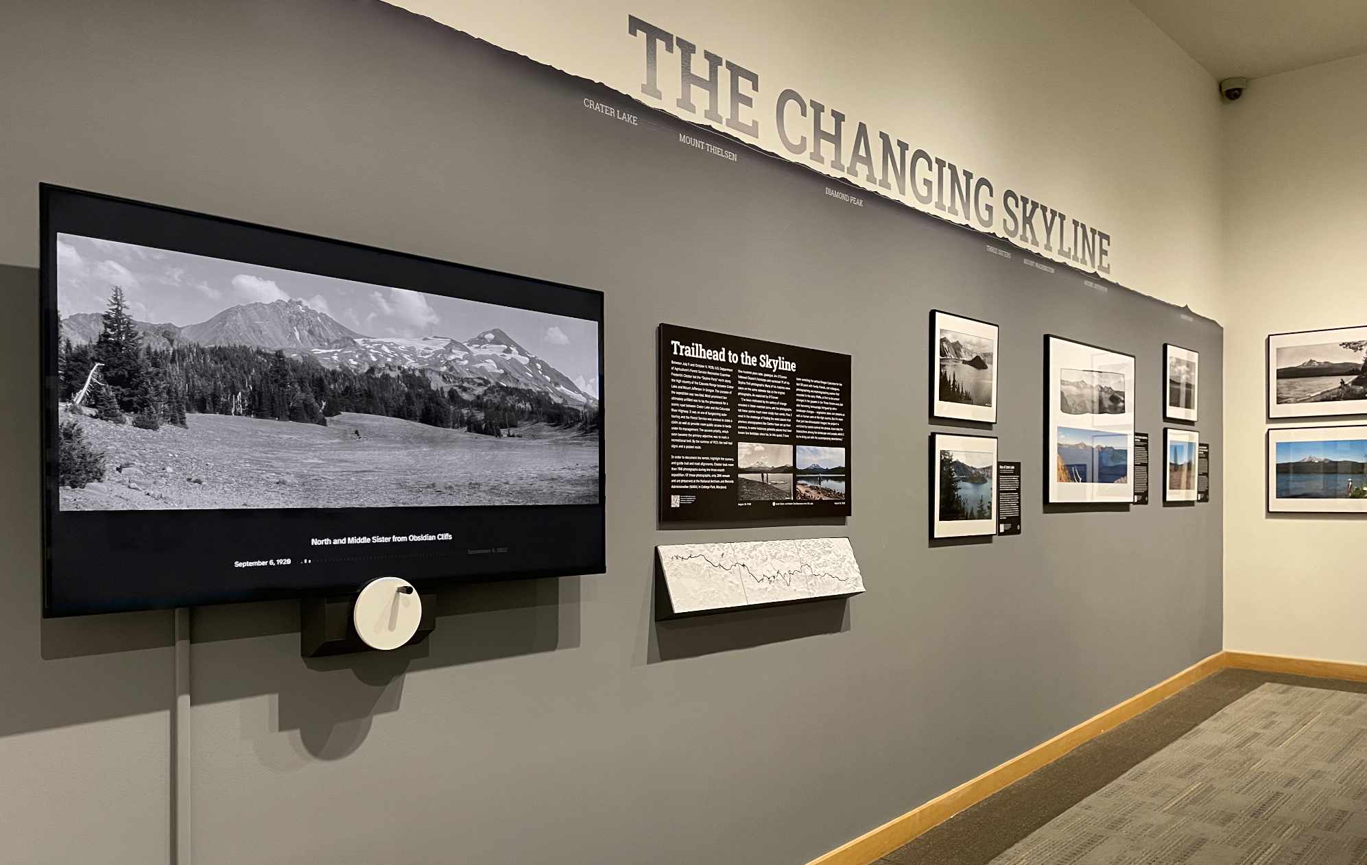 A narrow gallery under “The Changing Skyline” lettering: a wide screen showing a black-and-white mountain panorama, framed photographs along the wall, and the tactile topographic map on a low plinth.