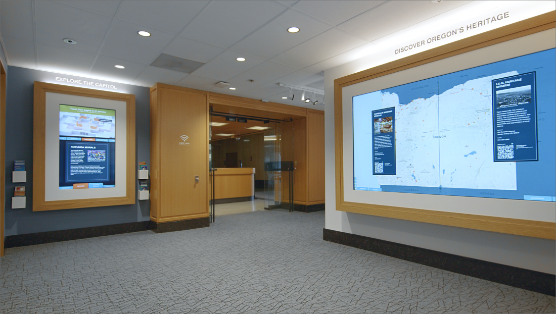 The Welcome Center interior: wood-framed touchscreens set into the wall beneath “Oregon’s Heritage” lettering, a staffed desk at the centre, and a large map screen to the right.